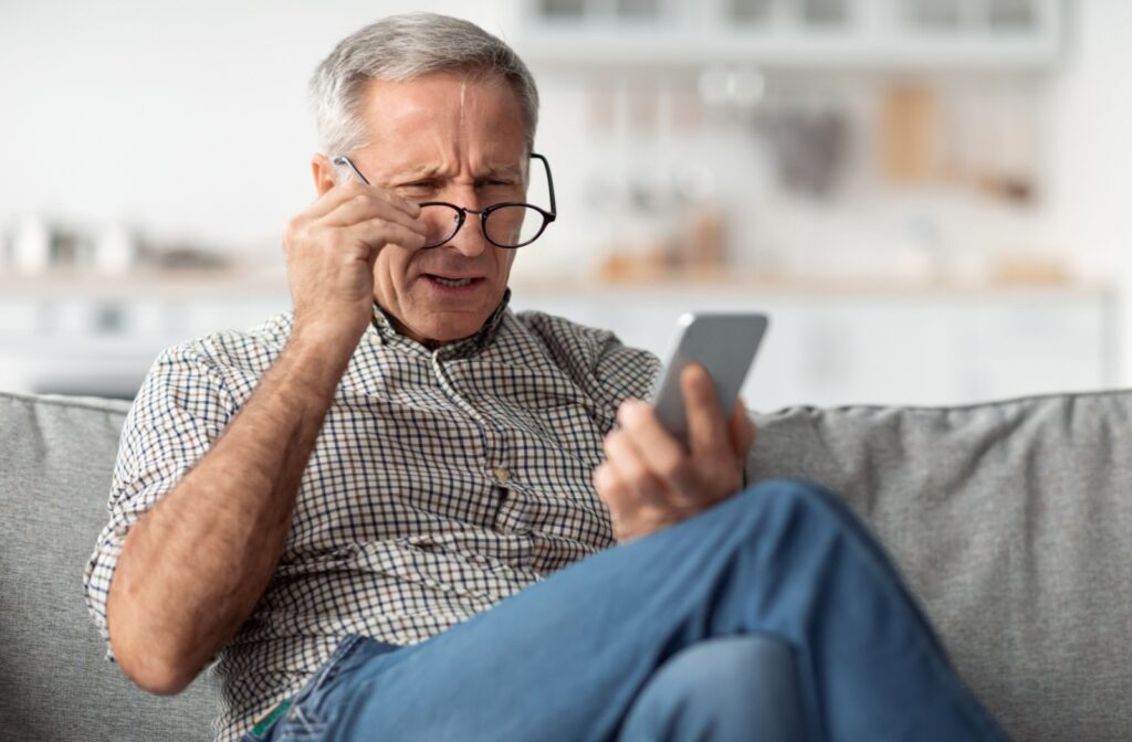 An older adult sitting on the couch, holding their glasses and squinting at their phone to see, wondering if their eyesight is getting worse.